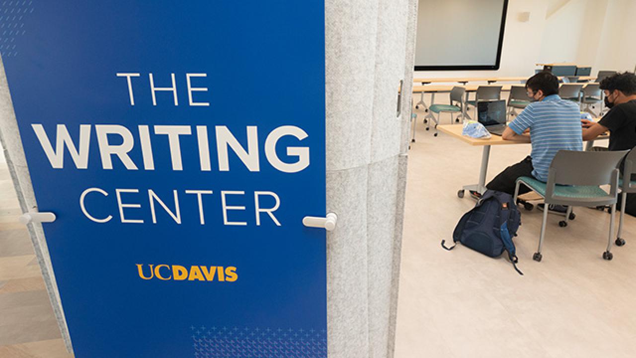 An image of the Writing Center sign on a panel in the facility with two students working at a table behind it