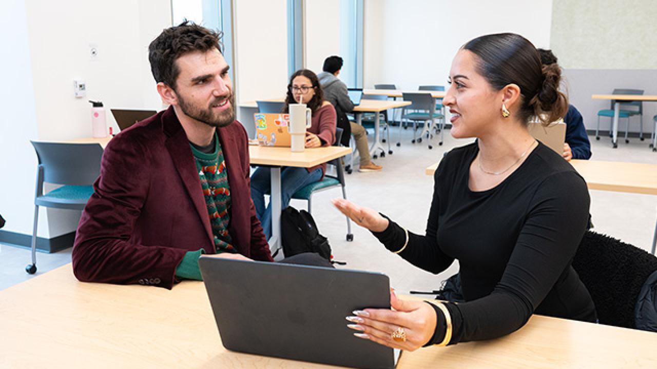 Photo of grad assistant and student seated at a large table looking at a laptop together with other students working at tables behind them.