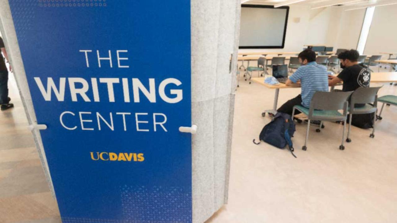 The photo features a 8-foot-tall blue sign attached toa wall that says Writing Center. Beside the wall is an opening into a classroom in the Writing Center. The viewer can see several long work tables with two people working on laptops seated next to each other at the nearest table.