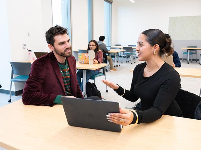 Photo of grad assistant and student seated at a large table looking at a laptop together with other students working at tables behind them.
