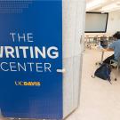 An image of the Writing Center sign on a panel in the facility with two students working at a table behind it