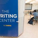 The photo features a 8-foot-tall blue sign attached toa wall that says Writing Center. Beside the wall is an opening into a classroom in the Writing Center. The viewer can see  several long work tables with two people working on laptops seated next to each other at the nearest table. 