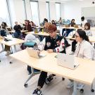 Writing Center large meeting area with students working in pairs at several long tables. 